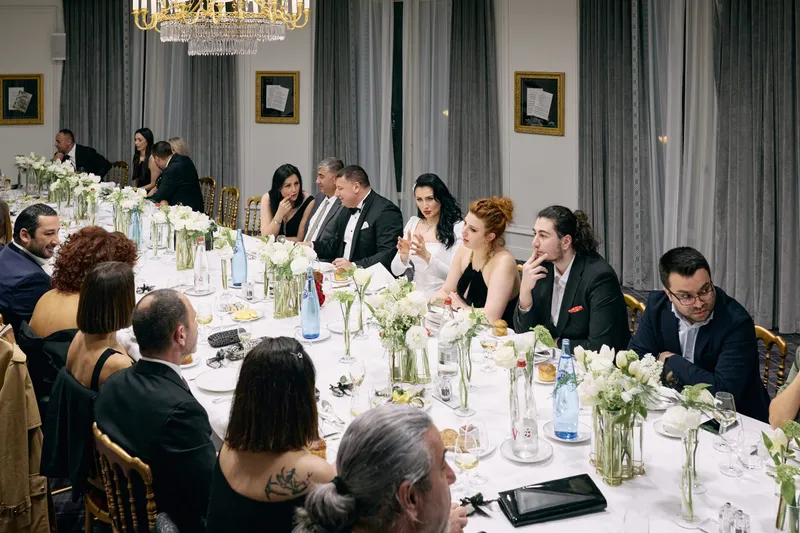 Guests seated along a long dinner table with white flowers and candles, viewed from the end of the room.