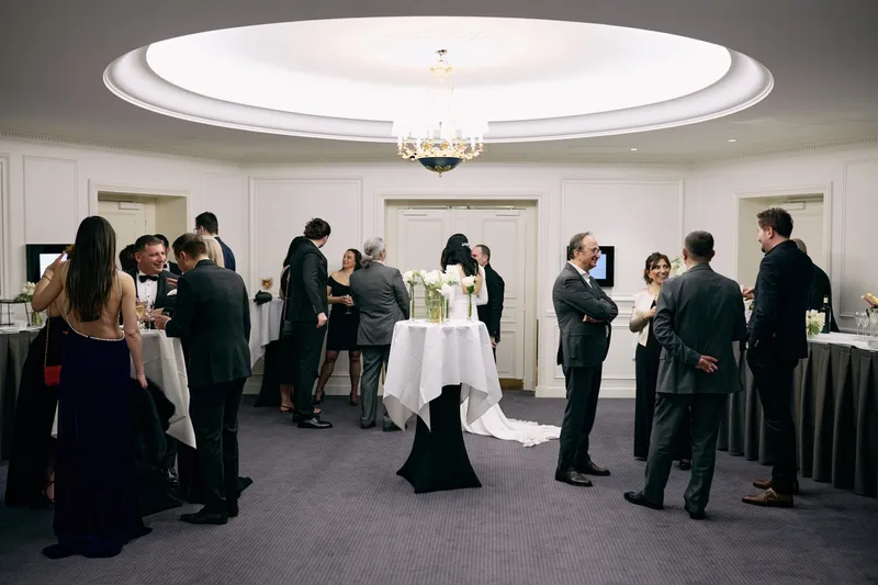 Guests in evening wear mingle with drinks in a hotel reception room under a domed ceiling.