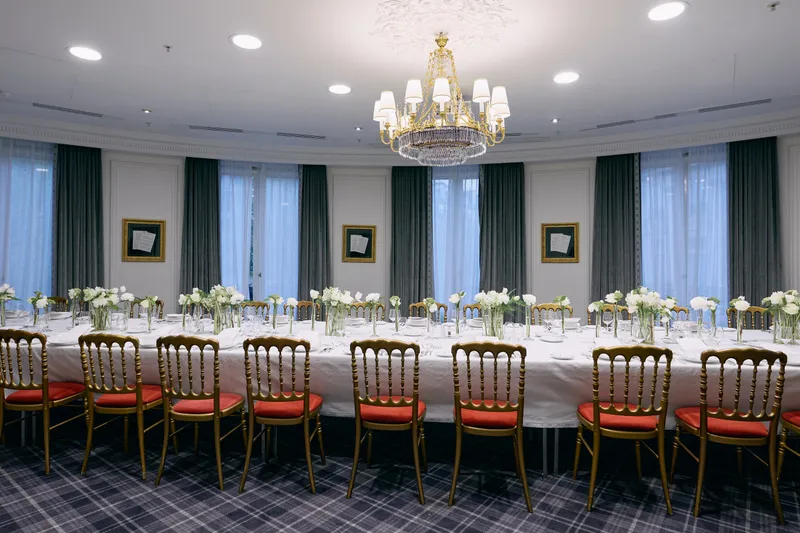A long reception table set with white linen, gold chairs and small white flower arrangements under a crystal chandelier.