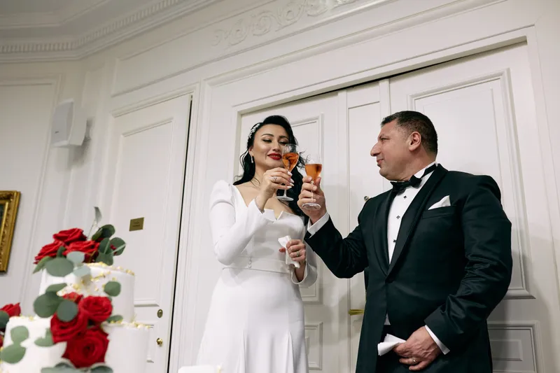 The bride and groom raise their glasses to each other in front of a red rose arrangement.